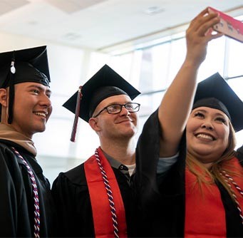 Three University of Phoenix graduates in regalia pose for a selfie at a commencement ceremony